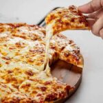 This photo shows a woman's hand removing a slice from a whole cheese pizza.